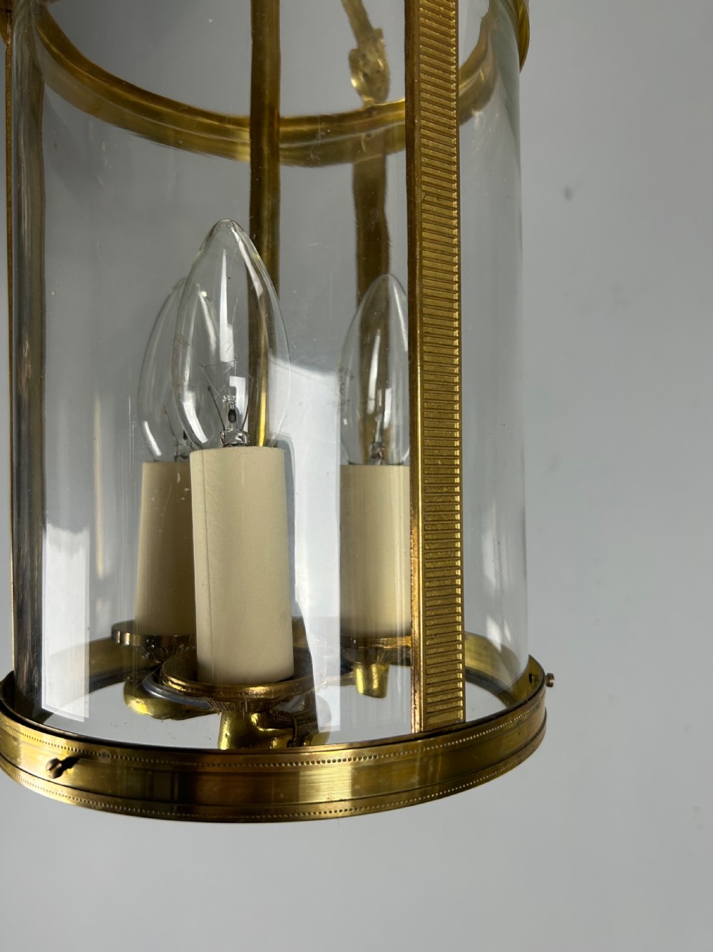 An antique French Empire style hanging brass lantern featuring a clear glass cylinder surrounding three candle-style light sockets and ornate scroll suspension arms. View IV