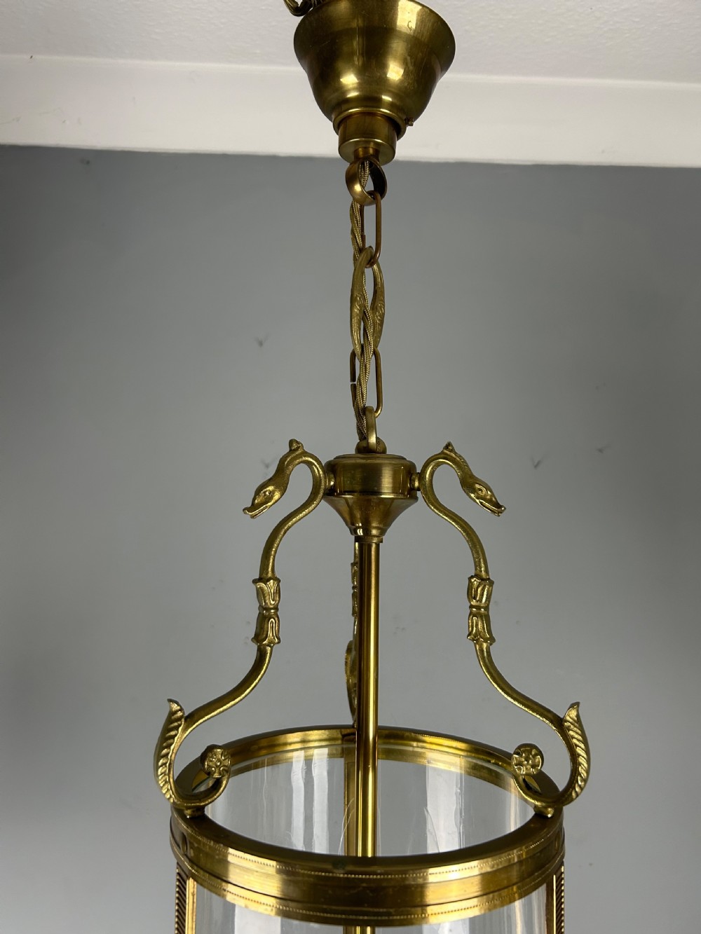 An antique French Empire style hanging brass lantern featuring a clear glass cylinder surrounding three candle-style light sockets and ornate scroll suspension arms. View V