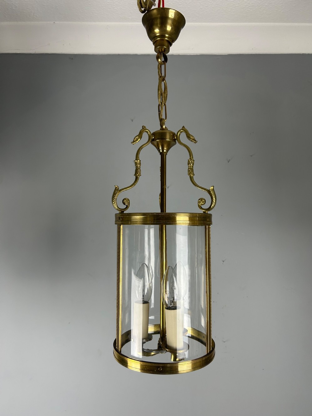 An antique French Empire style hanging brass lantern featuring a clear glass cylinder surrounding three candle-style light sockets and ornate scroll suspension arms. View VI
