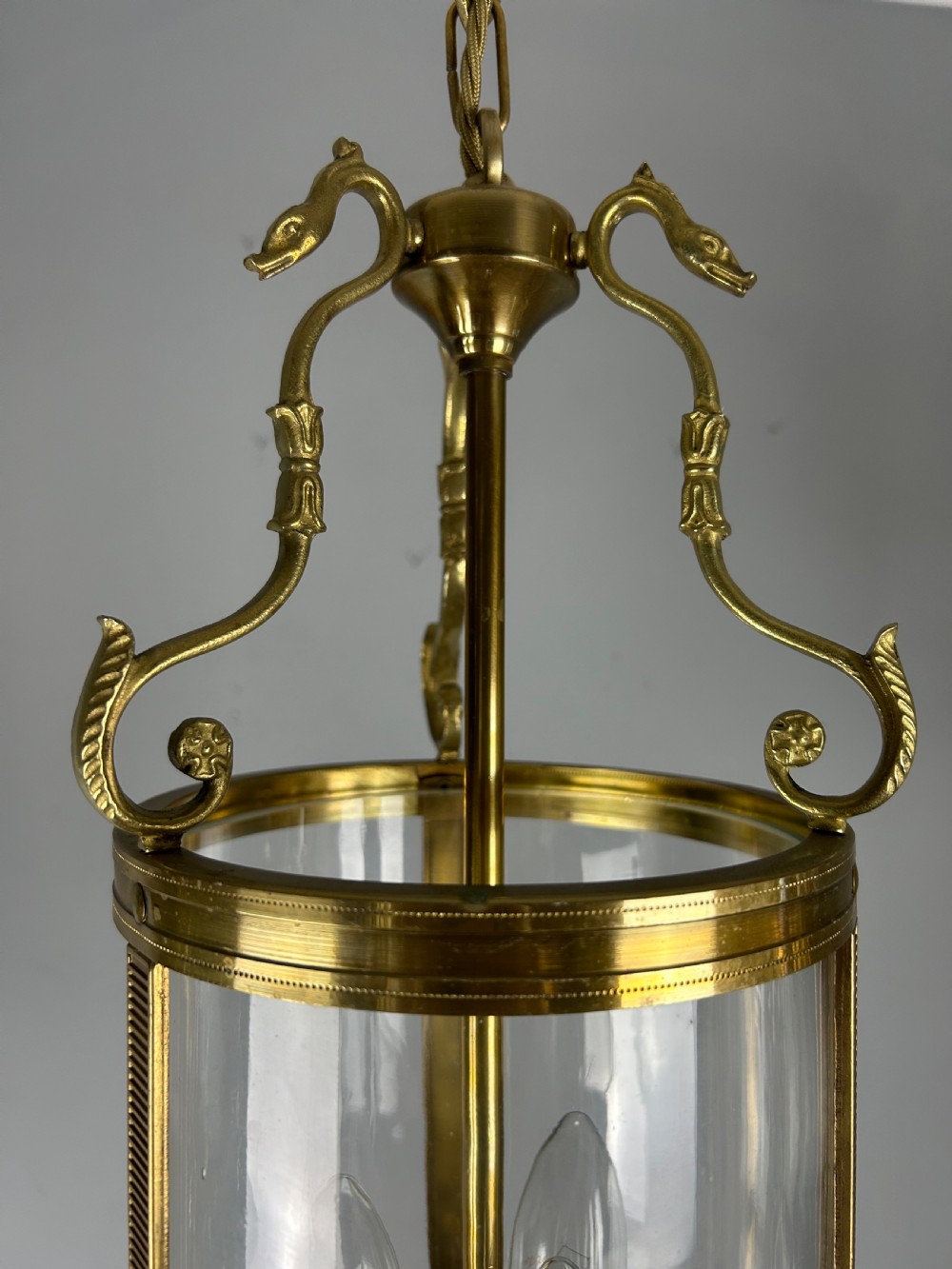 An antique French Empire style hanging brass lantern featuring a clear glass cylinder surrounding three candle-style light sockets and ornate scroll suspension arms. View VII