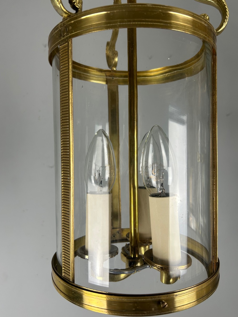 An antique French Empire style hanging brass lantern featuring a clear glass cylinder surrounding three candle-style light sockets and ornate scroll suspension arms. View VIII