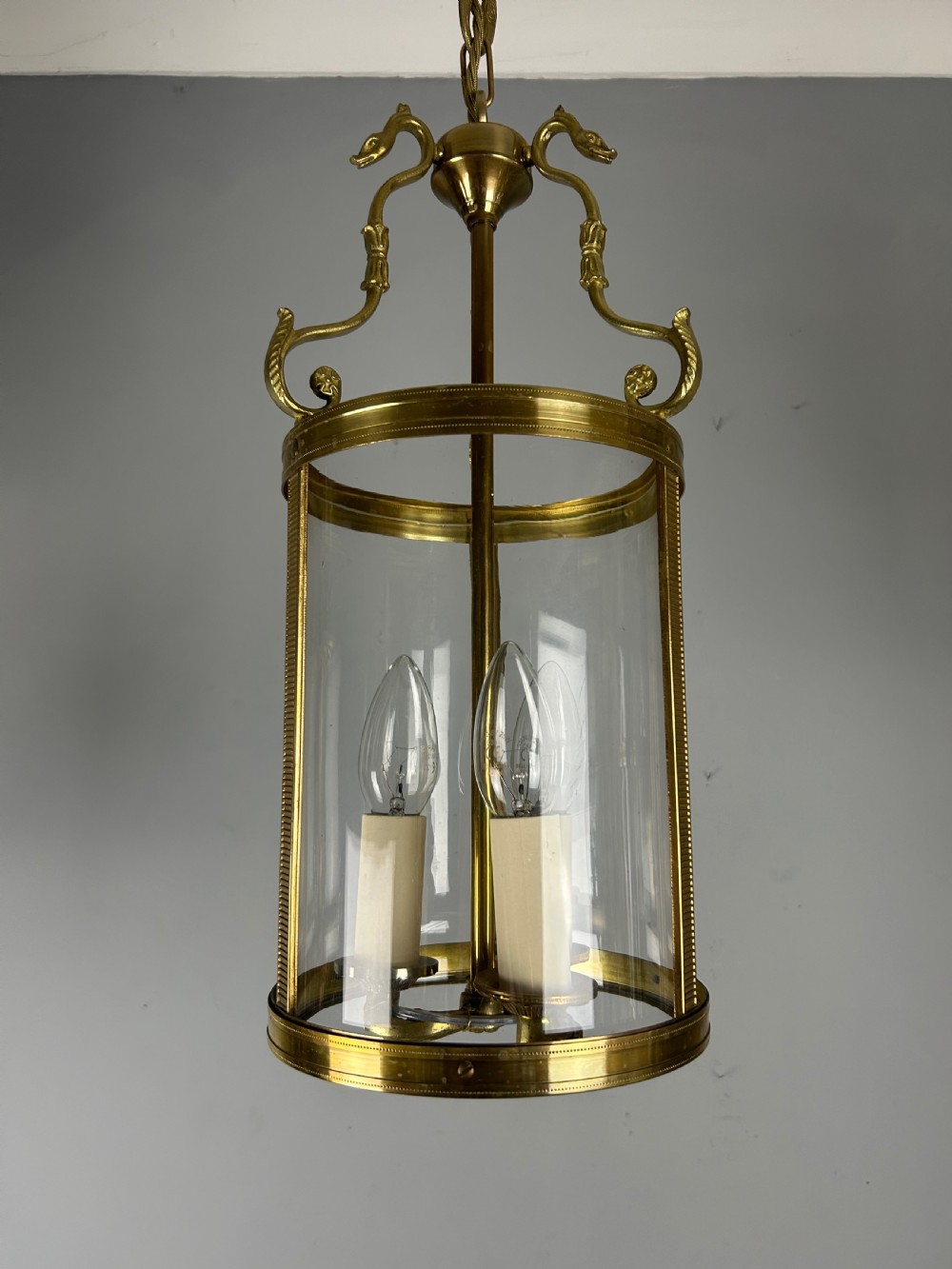 An antique French Empire style hanging brass lantern featuring a clear glass cylinder surrounding three candle-style light sockets and ornate scroll suspension arms. View IX