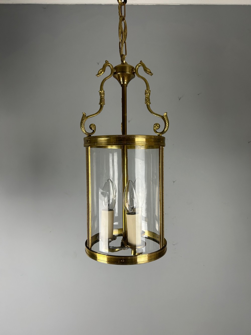 An antique French Empire style hanging brass lantern featuring a clear glass cylinder surrounding three candle-style light sockets and ornate scroll suspension arms. View X