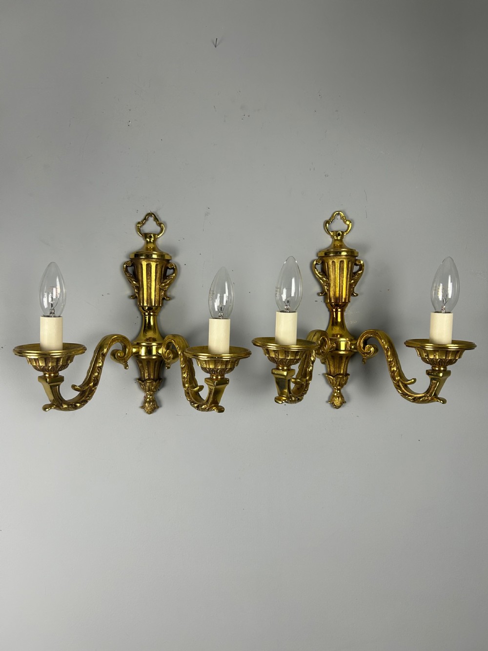 A pair of ornate French gilt brass wall lights featuring twin scrolling arms and central urn motifs against a neutral background.