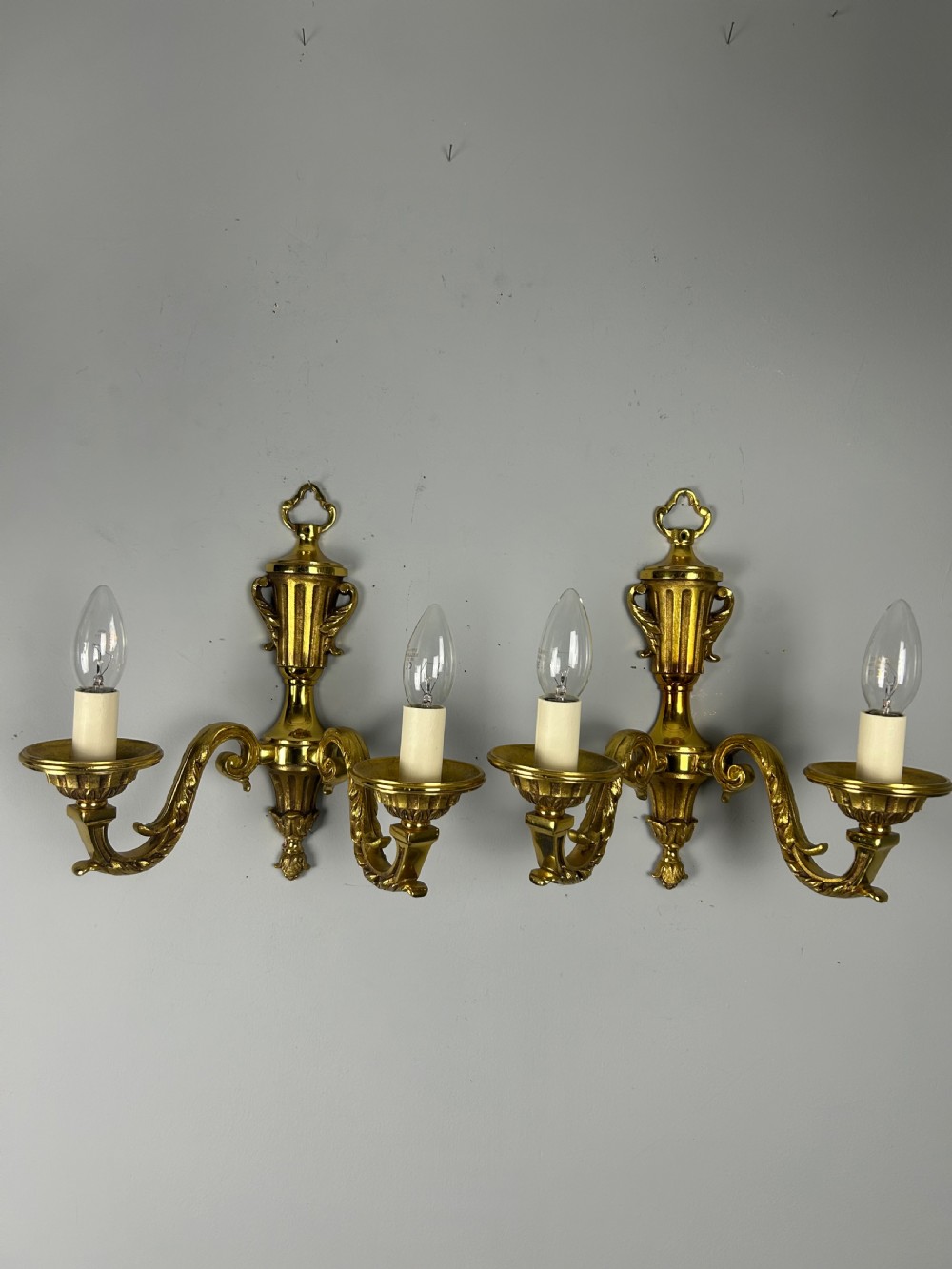A pair of ornate French gilt brass wall lights featuring twin scrolling arms and central urn motifs against a neutral background. View IV