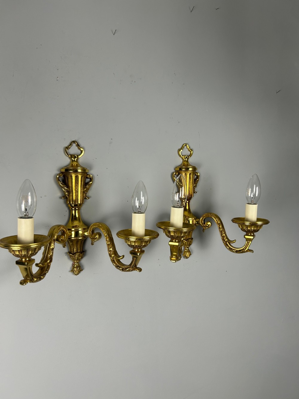 A pair of ornate French gilt brass wall lights featuring twin scrolling arms and central urn motifs against a neutral background. View V