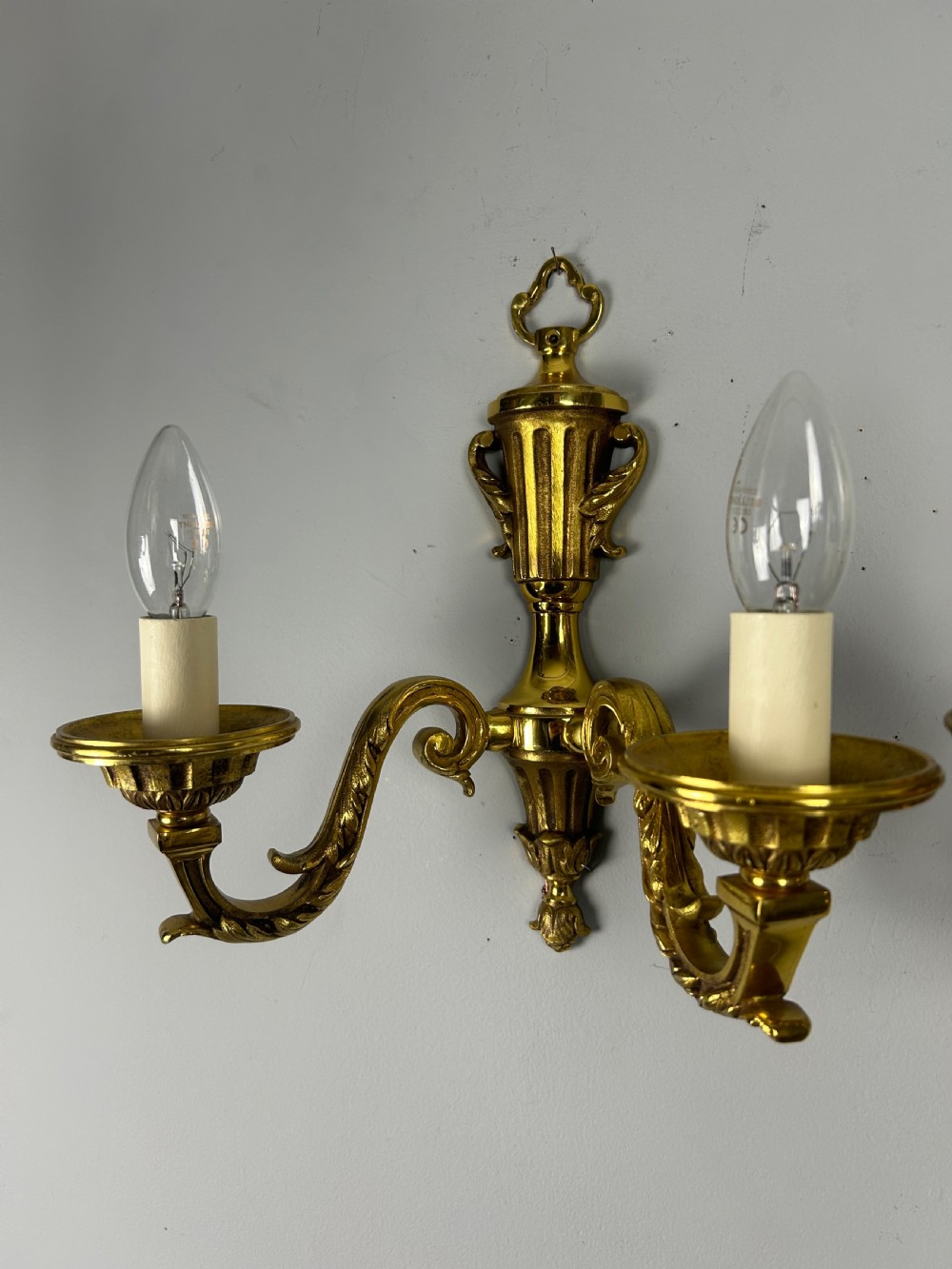 A pair of ornate French gilt brass wall lights featuring twin scrolling arms and central urn motifs against a neutral background. View VII