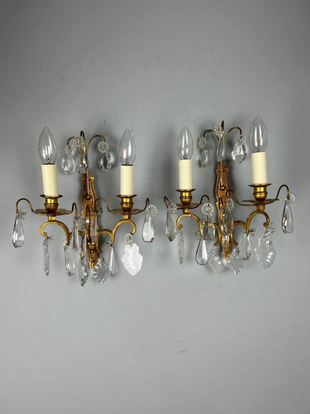 A pair of antique twin-arm wall lights featuring gilded frames and cascading crystal drops against a neutral background. View II