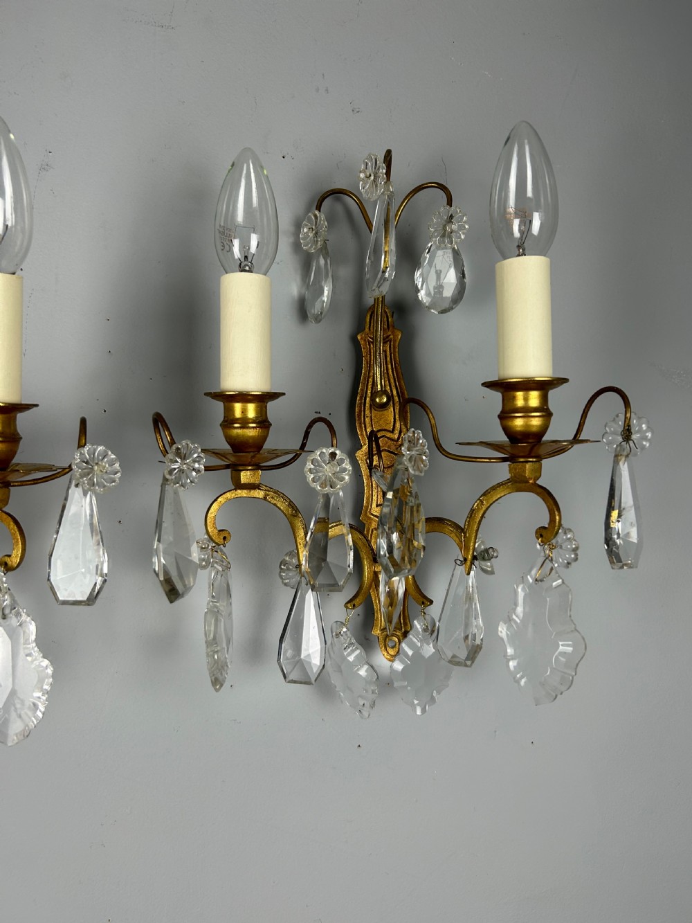 A pair of antique twin-arm wall lights featuring gilded frames and cascading crystal drops against a neutral background. View V