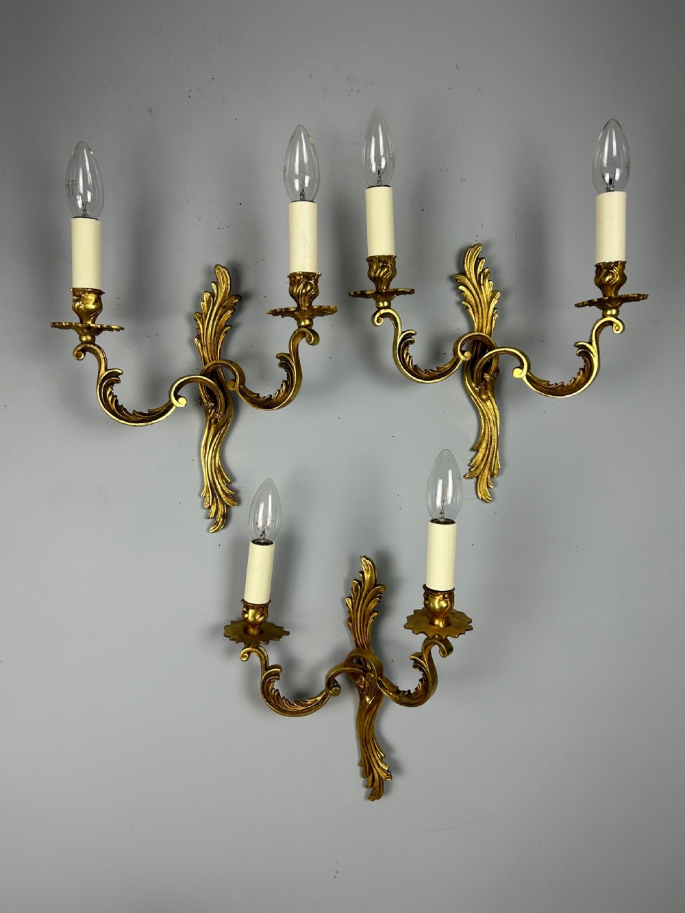 A set of three French gilt metal wall sconces featuring scrolled arms and decorative acanthus leaf backplates, fitted with candle-style bulbs.