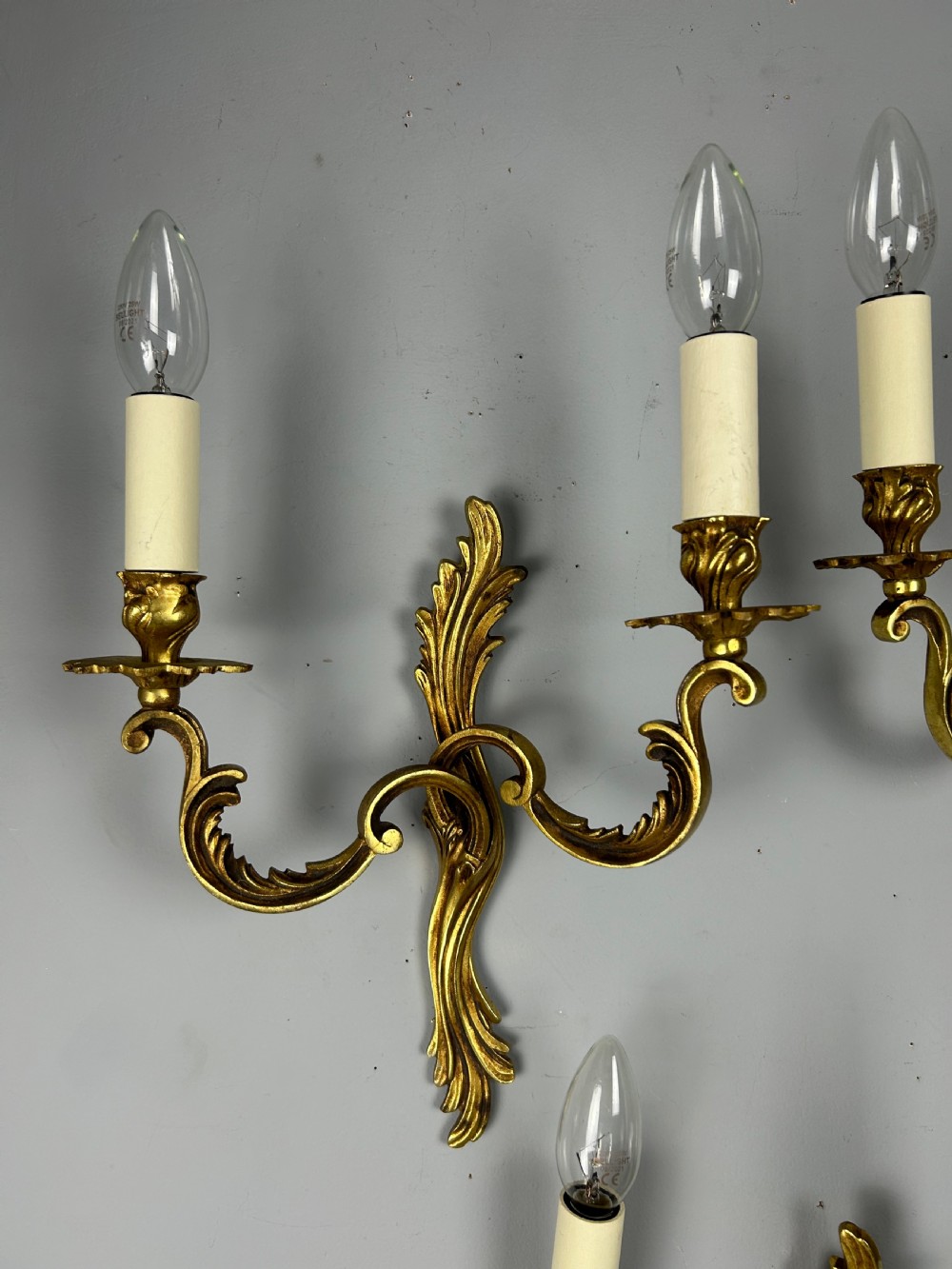 A set of three French gilt metal wall sconces featuring scrolled arms and decorative acanthus leaf backplates, fitted with candle-style bulbs. View V