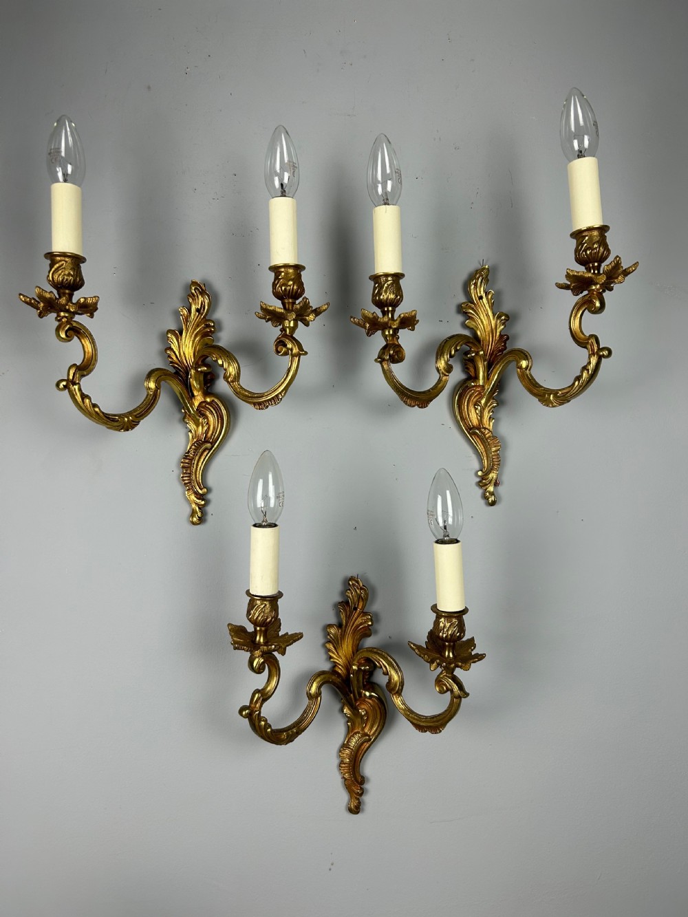 A set of three antique French gilded twin arm wall lights mounted against a neutral grey wall.
