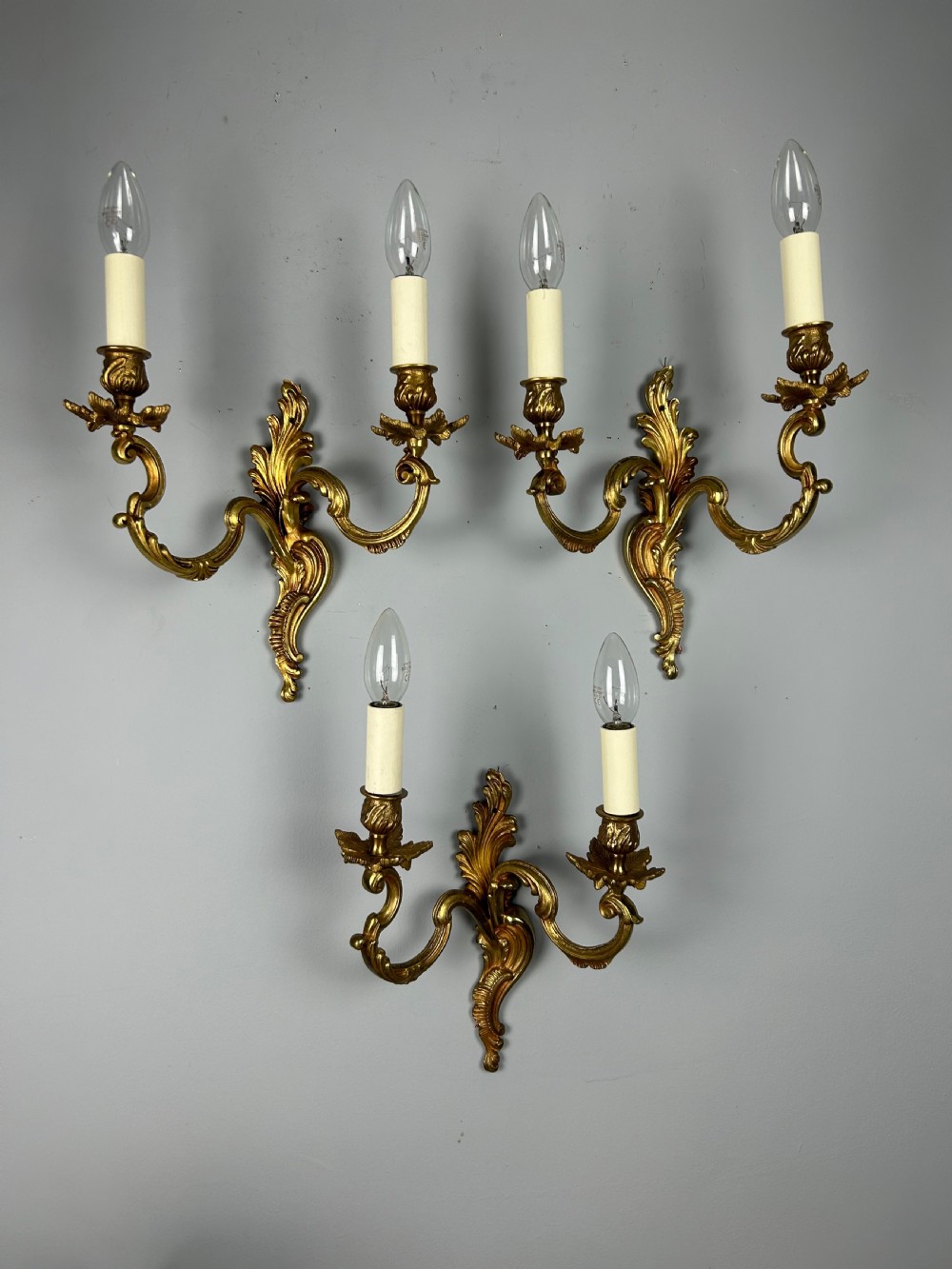 A set of three antique French gilded twin arm wall lights mounted against a neutral grey wall. View II