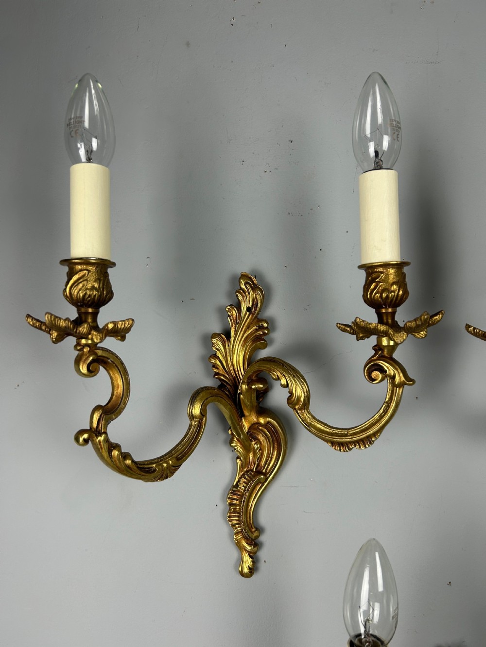 A set of three antique French gilded twin arm wall lights mounted against a neutral grey wall. View IX