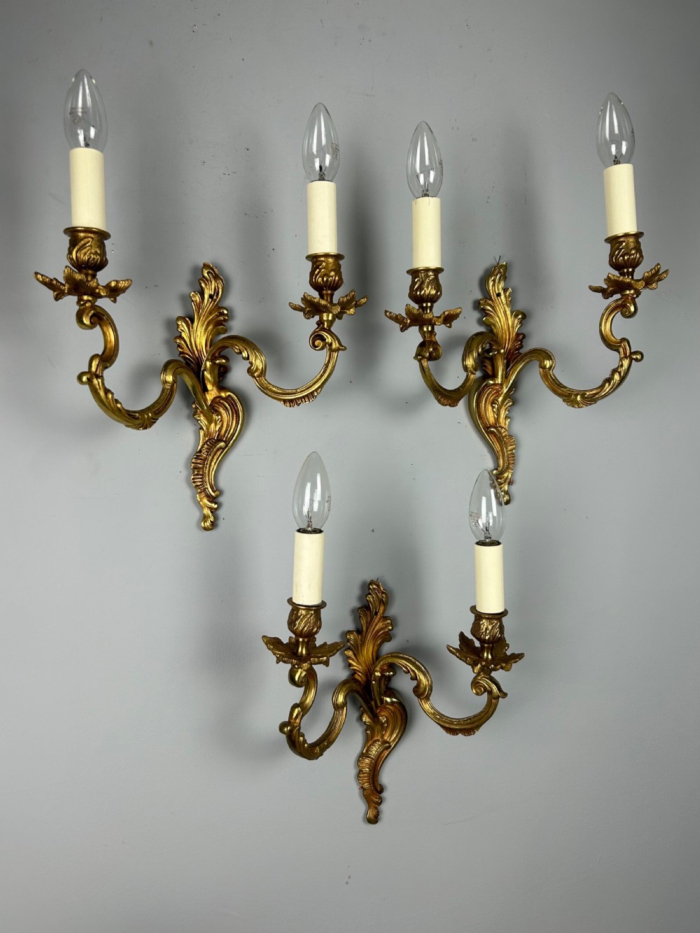 A set of three antique French gilded twin arm wall lights mounted against a neutral grey wall. View X