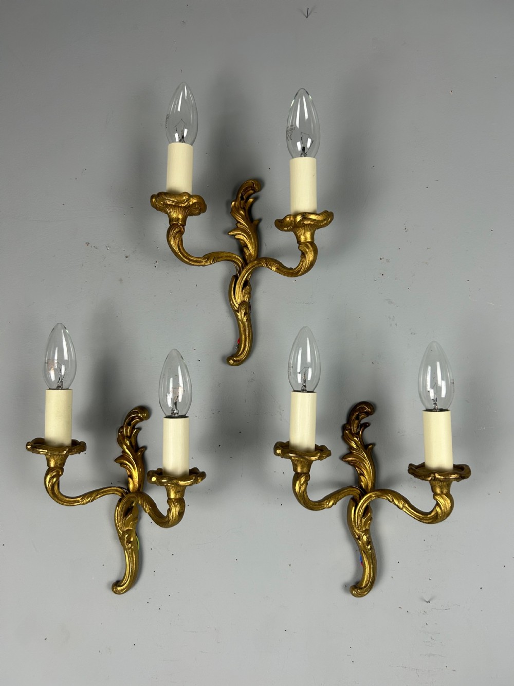 Set of three French gilt brass two-arm wall sconces featuring leaf ornamentation and simulated candle sockets.