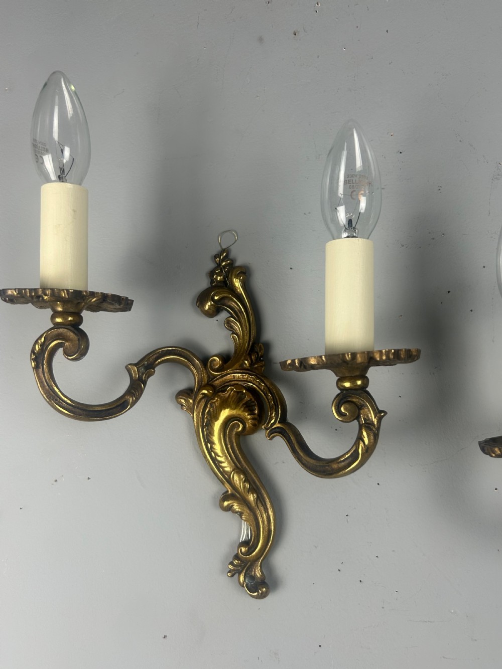A pair of French Rococo style gilded brass twin-arm wall lights with foliate scrollwork and white candle sleeves. View II