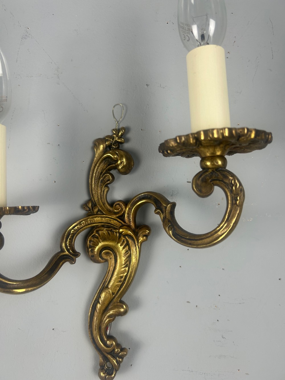 A pair of French Rococo style gilded brass twin-arm wall lights with foliate scrollwork and white candle sleeves. View III