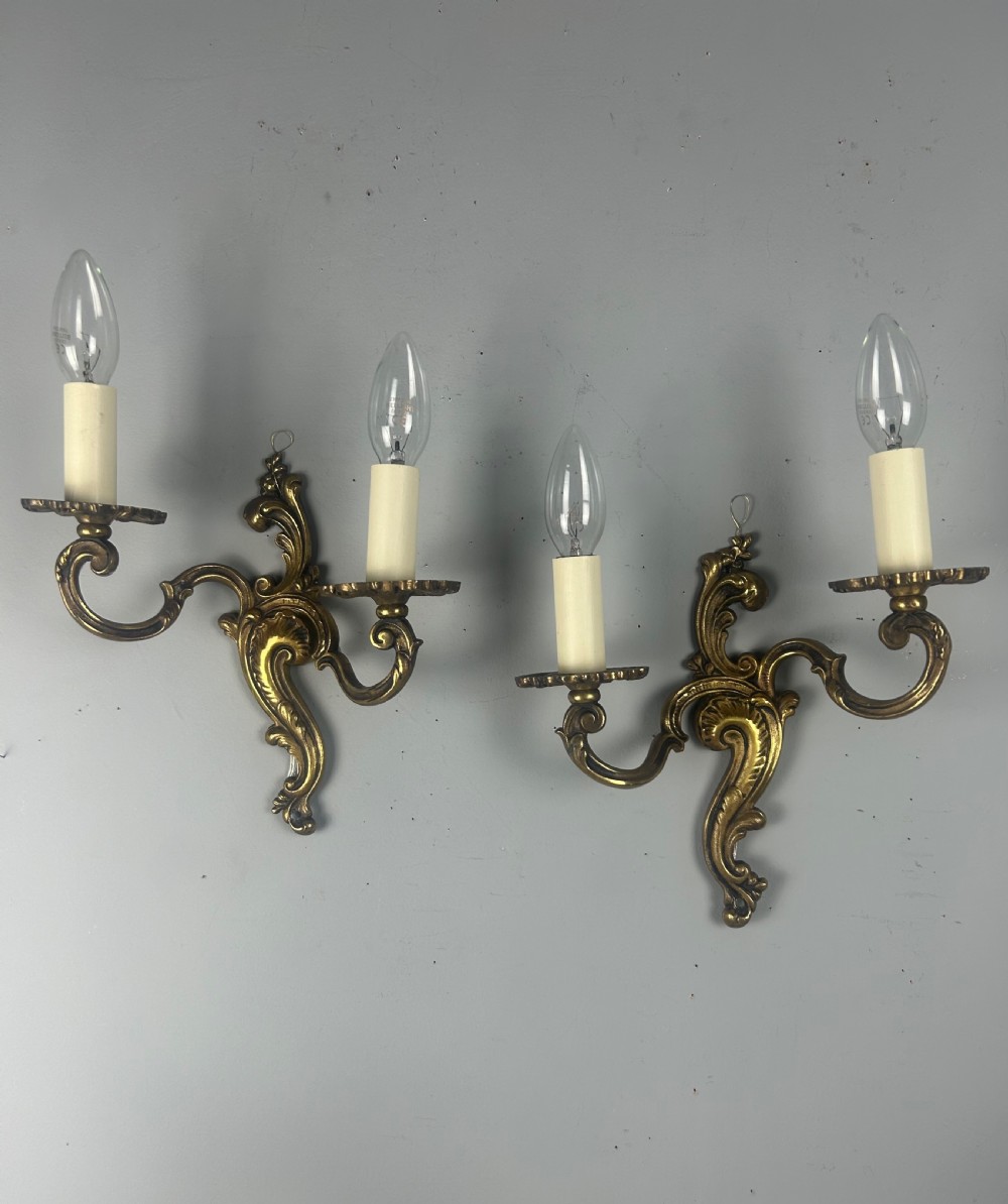 A pair of French Rococo style gilded brass twin-arm wall lights with foliate scrollwork and white candle sleeves. View IV