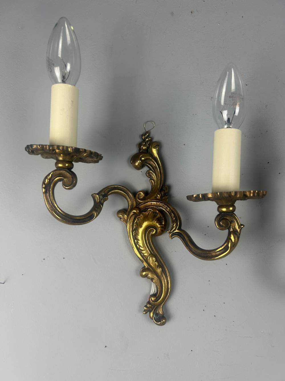 A pair of French Rococo style gilded brass twin-arm wall lights with foliate scrollwork and white candle sleeves. View V