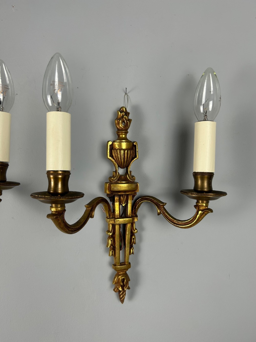 A pair of French Louis XVI style two-arm gilded metal wall lights featuring classical urn finials, tapered backplates, and faux candle sleeves. View II