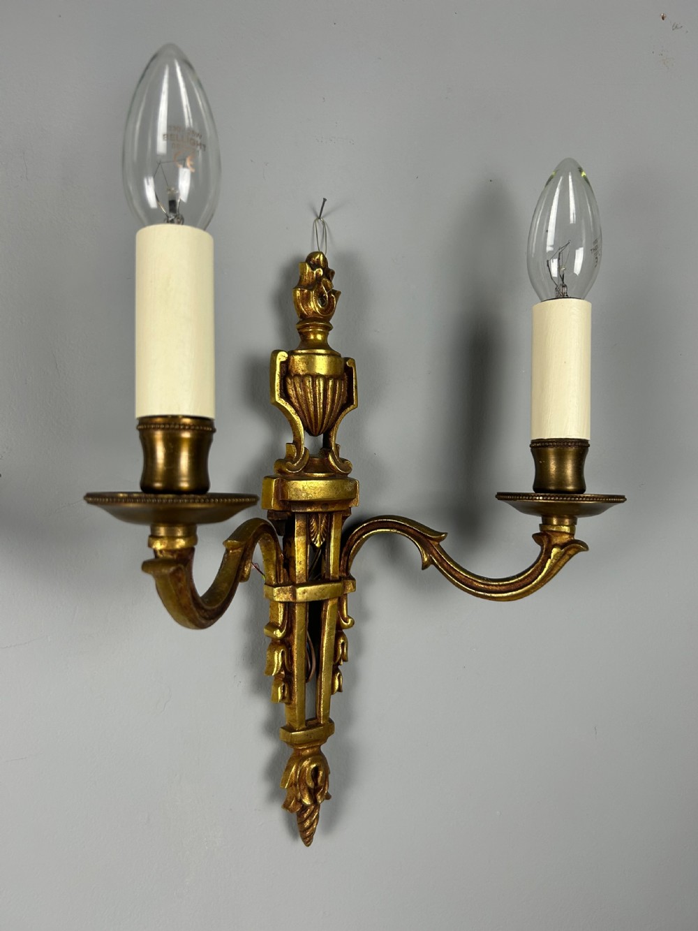 A pair of French Louis XVI style two-arm gilded metal wall lights featuring classical urn finials, tapered backplates, and faux candle sleeves. View III