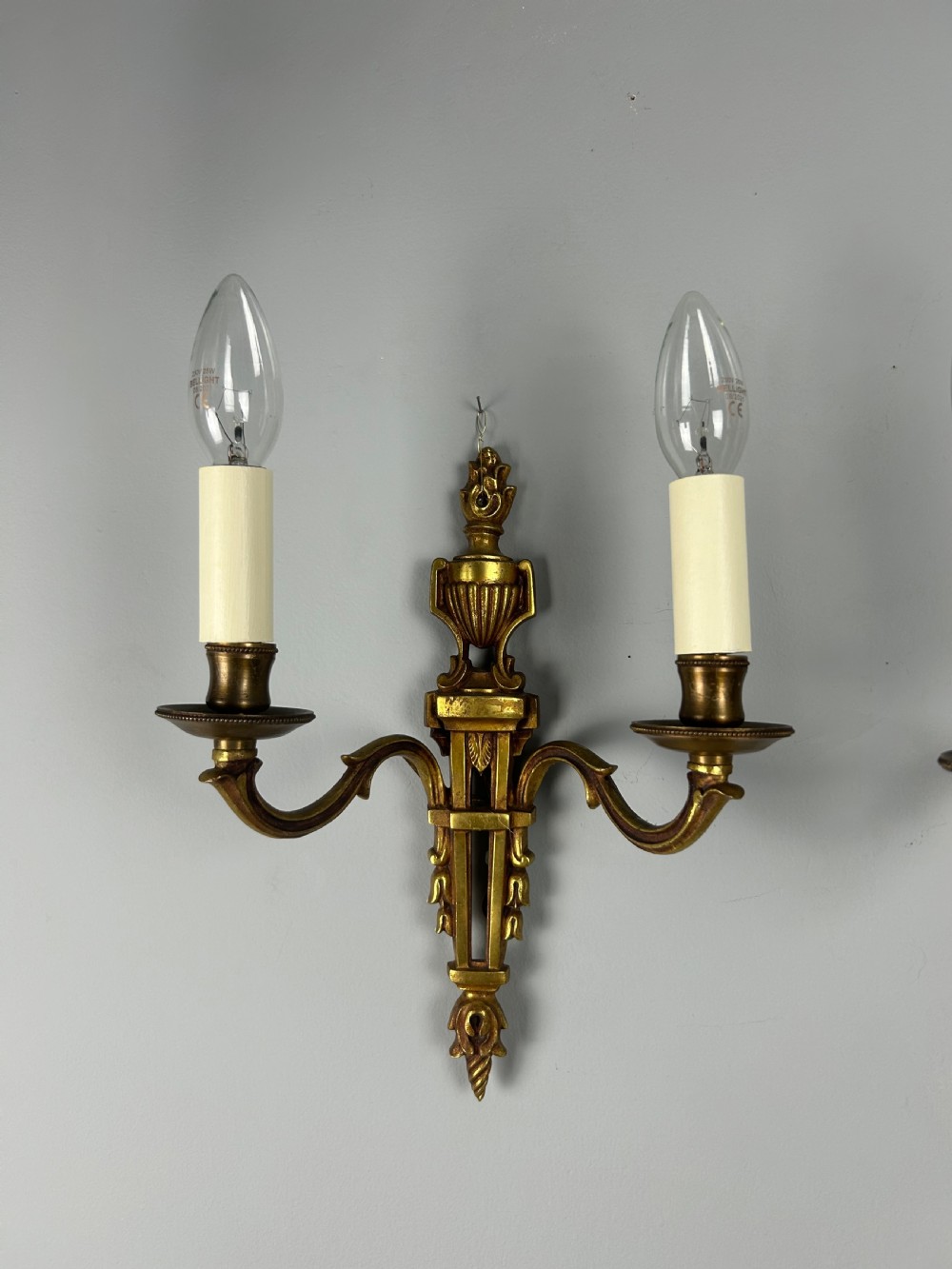 A pair of French Louis XVI style two-arm gilded metal wall lights featuring classical urn finials, tapered backplates, and faux candle sleeves. View IV