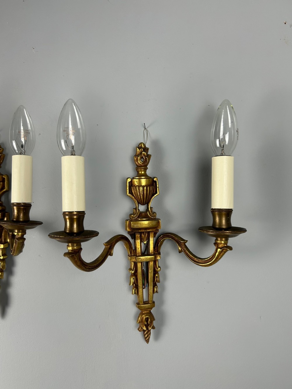 A pair of French Louis XVI style two-arm gilded metal wall lights featuring classical urn finials, tapered backplates, and faux candle sleeves. View V