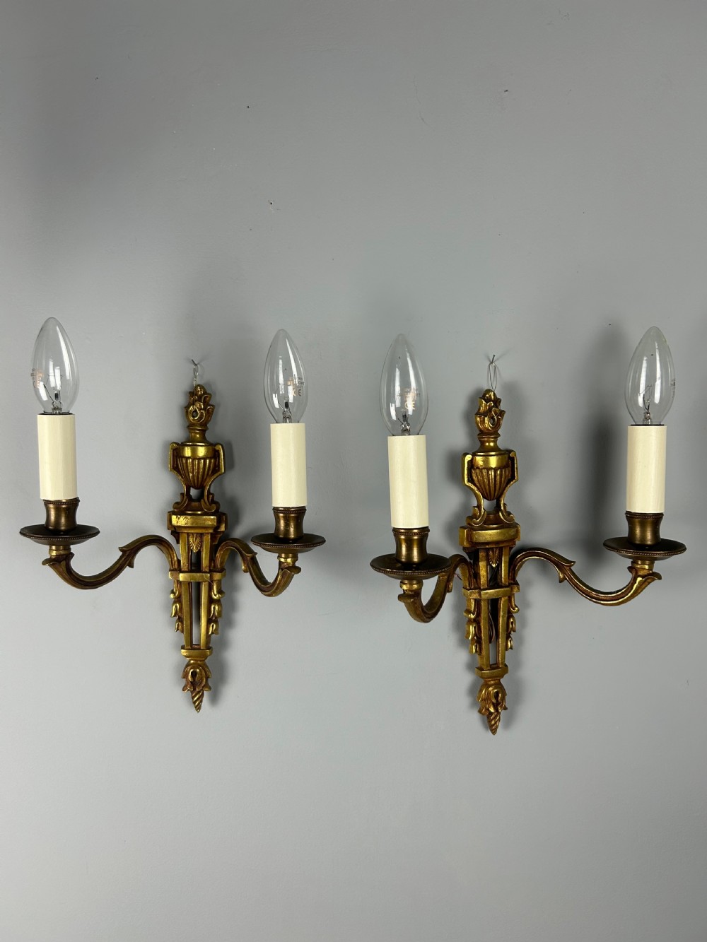 A pair of French Louis XVI style two-arm gilded metal wall lights featuring classical urn finials, tapered backplates, and faux candle sleeves. View VI