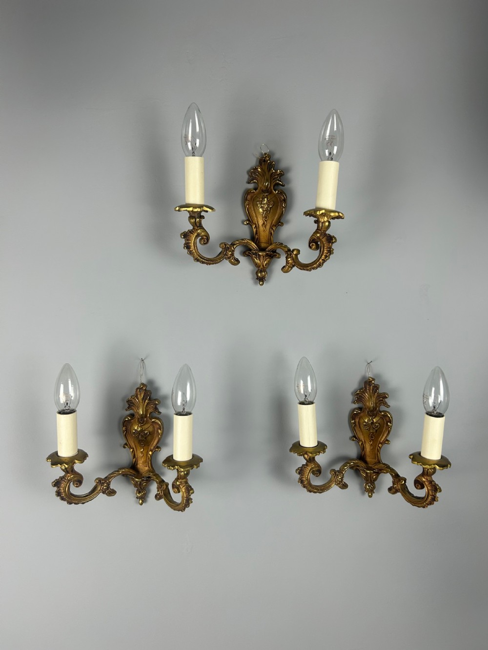 A set of three French Rococo style gilded brass wall lights with twin scroll arms and candle-shaped bulb holders.