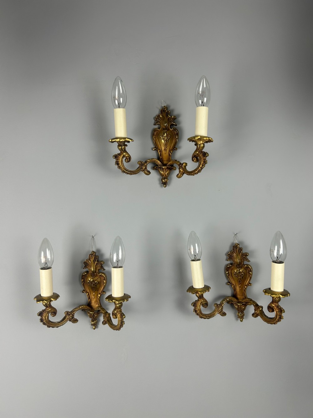 A set of three French Rococo style gilded brass wall lights with twin scroll arms and candle-shaped bulb holders. View VIII