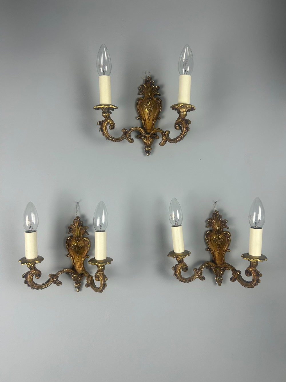 A set of three French Rococo style gilded brass wall lights with twin scroll arms and candle-shaped bulb holders. View X