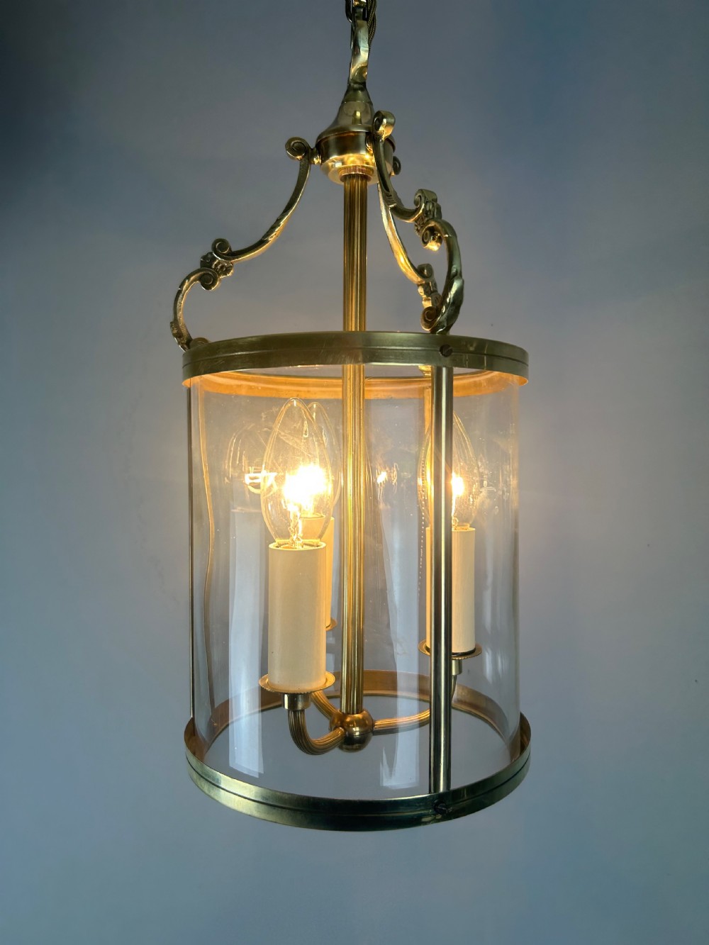 A polished brass cylindrical pendant lantern with clear curved glass panels housing a three-light candle cluster inside. View II