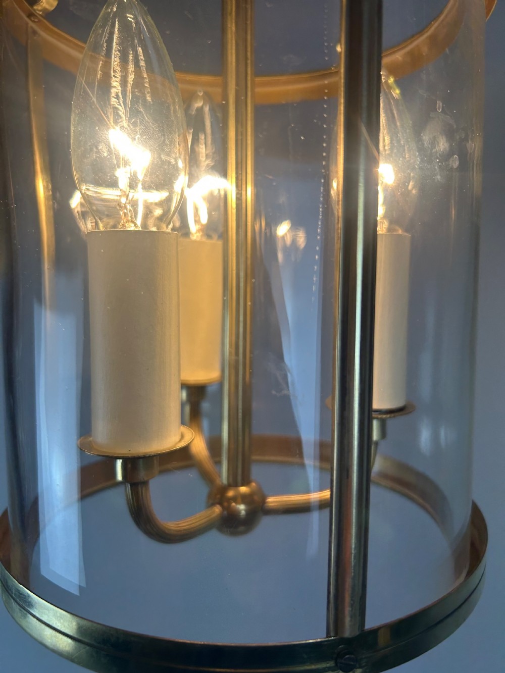 A polished brass cylindrical pendant lantern with clear curved glass panels housing a three-light candle cluster inside. View III