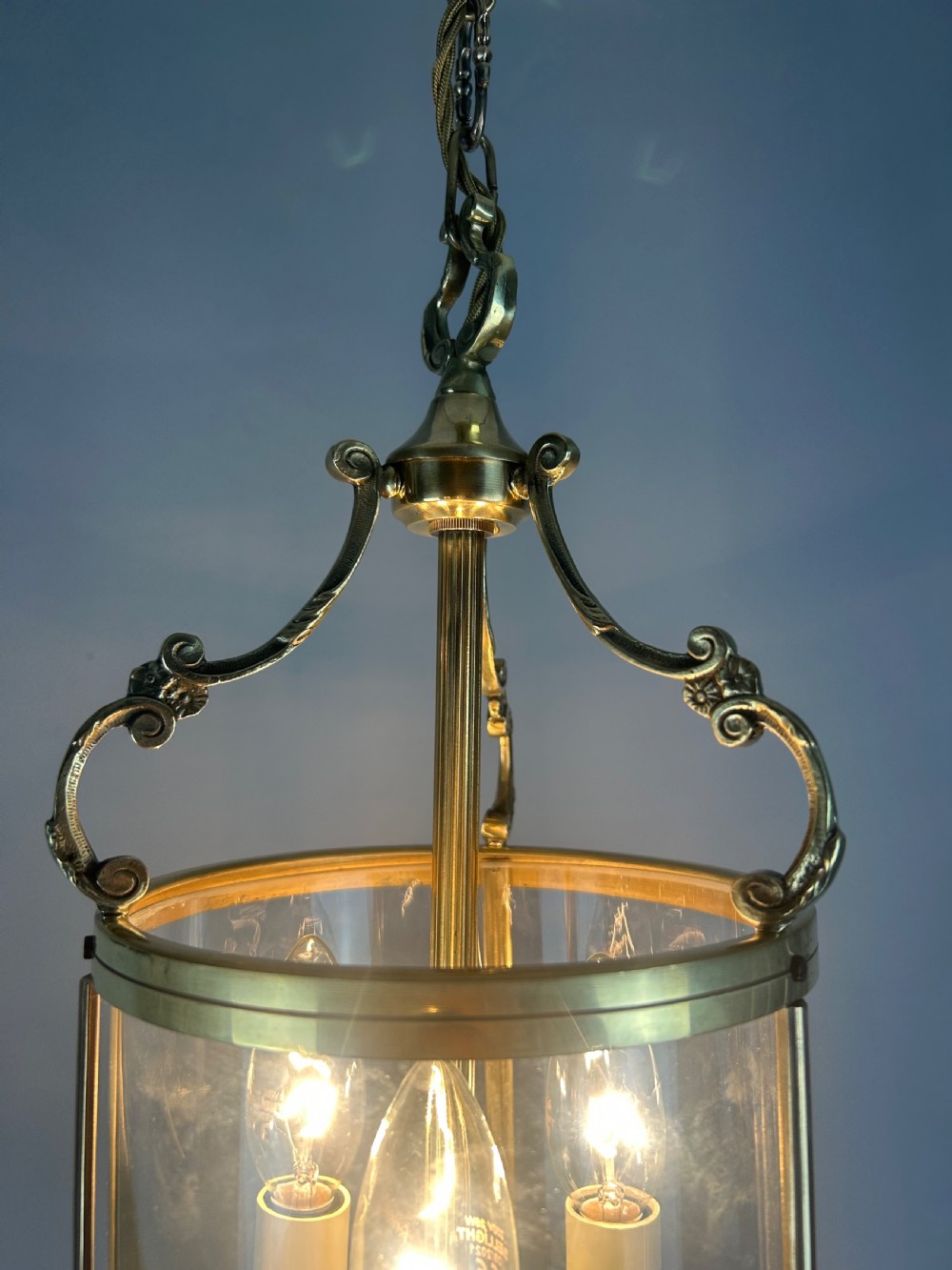 A polished brass cylindrical pendant lantern with clear curved glass panels housing a three-light candle cluster inside. View V