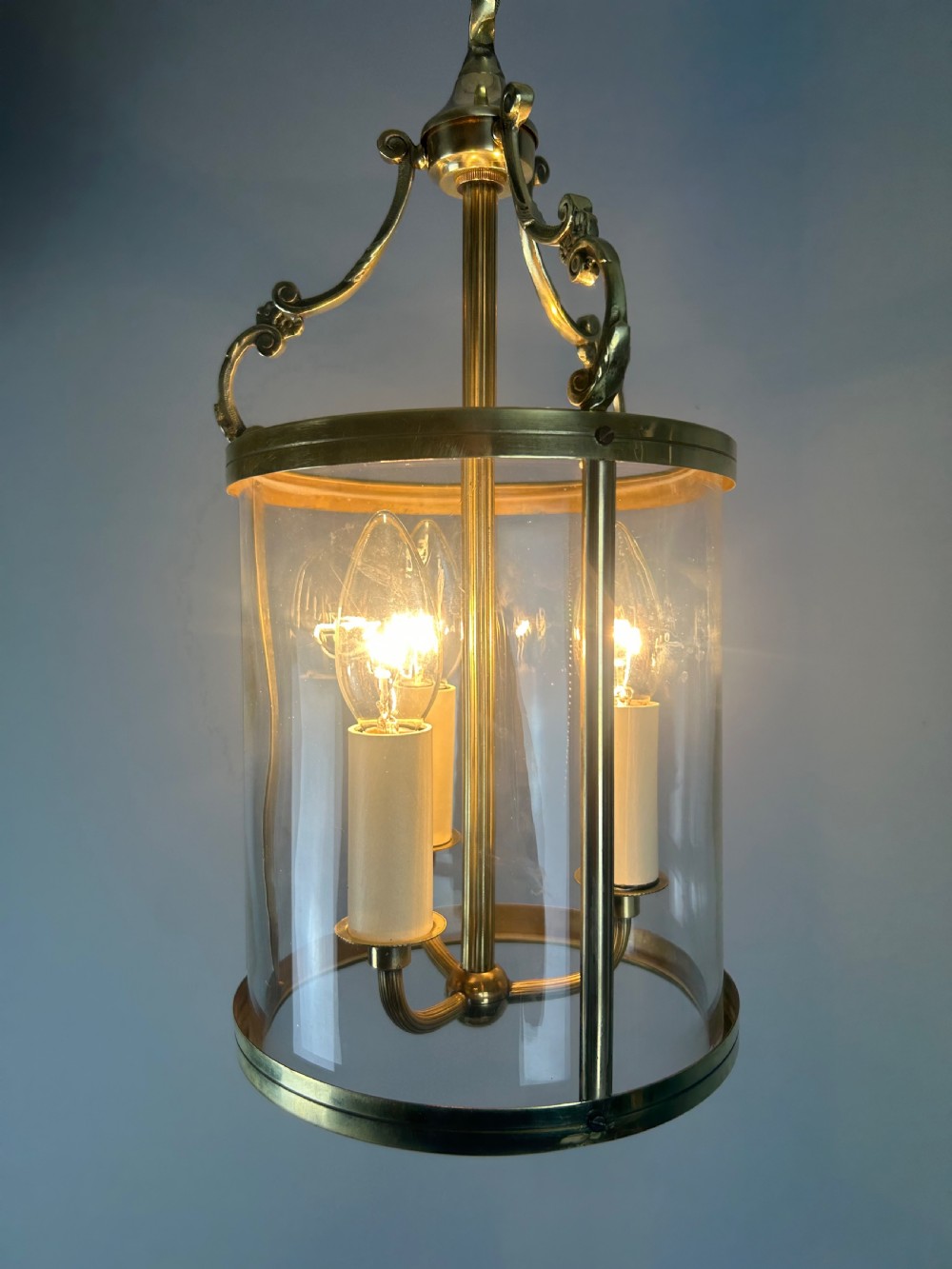 A polished brass cylindrical pendant lantern with clear curved glass panels housing a three-light candle cluster inside. View VI
