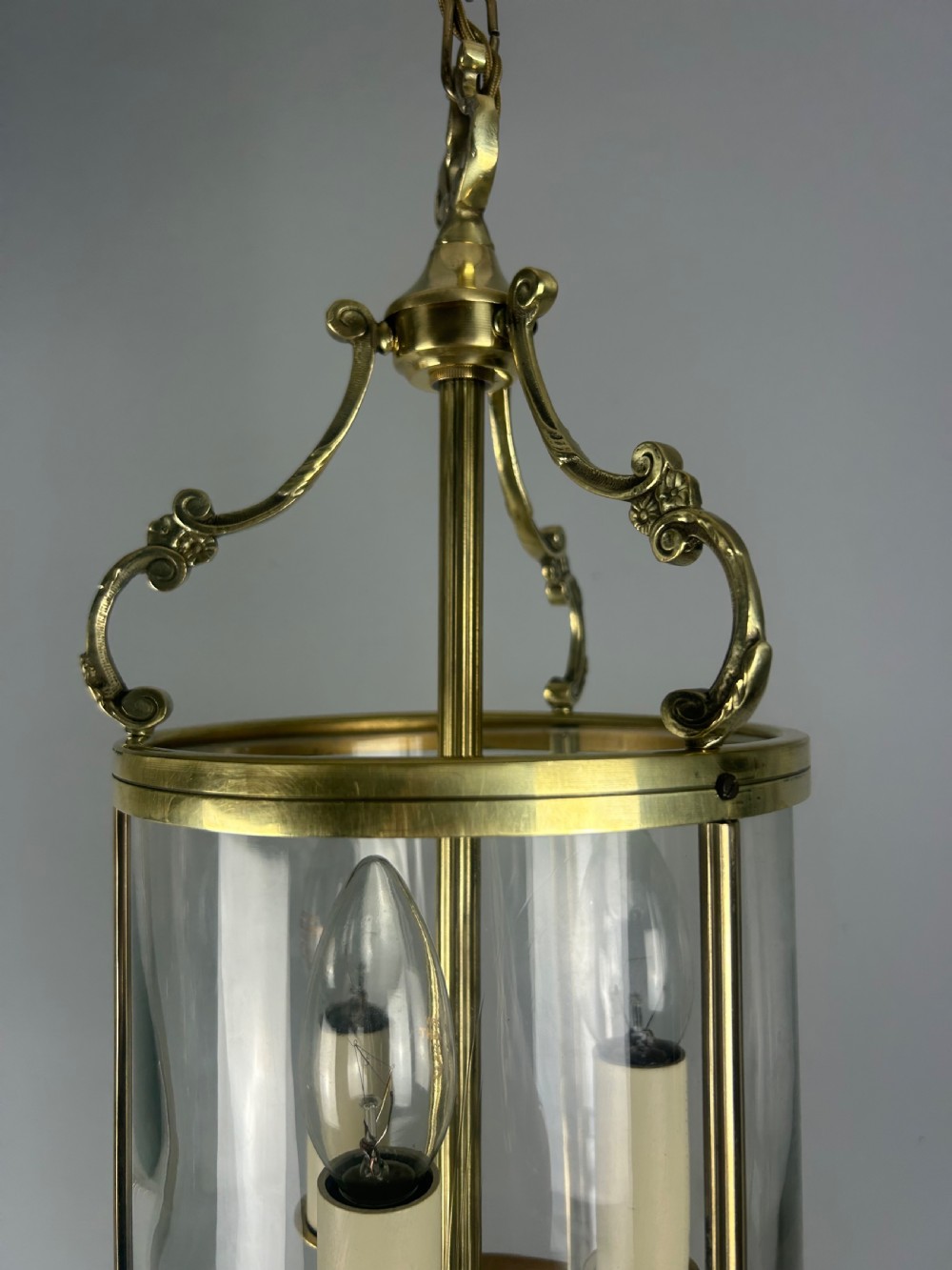 A polished brass cylindrical pendant lantern with clear curved glass panels housing a three-light candle cluster inside. View VII