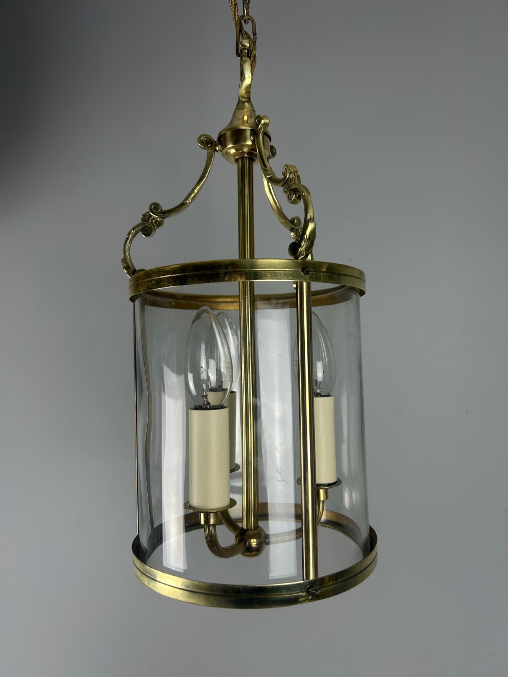 A polished brass cylindrical pendant lantern with clear curved glass panels housing a three-light candle cluster inside. View X