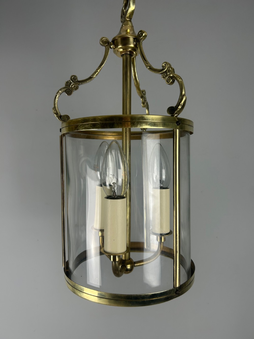 A polished brass cylindrical pendant lantern with clear curved glass panels housing a three-light candle cluster inside. View XI