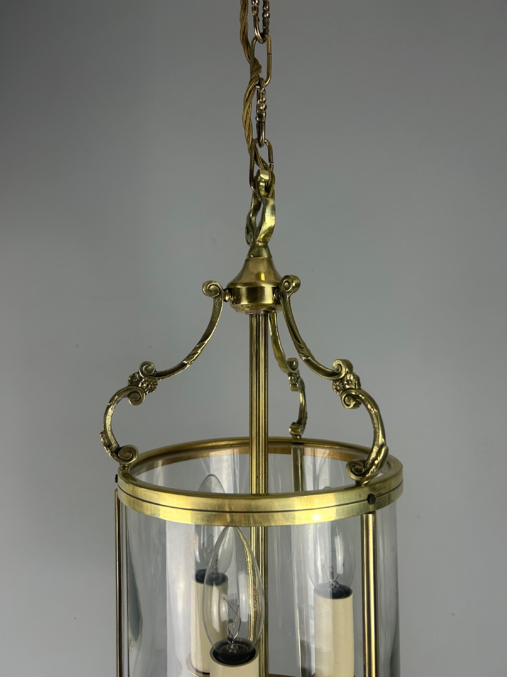A polished brass cylindrical pendant lantern with clear curved glass panels housing a three-light candle cluster inside. View XIII
