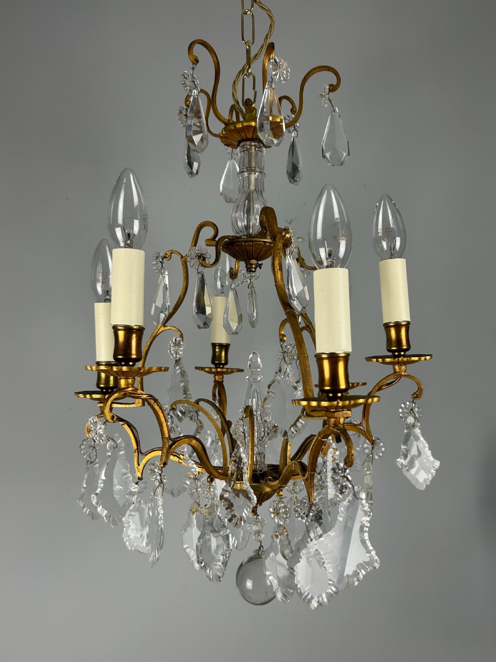 A French brass six-light chandelier featuring curved scroll arms, candle sleeves, and suspended faceted crystal glass droplets.