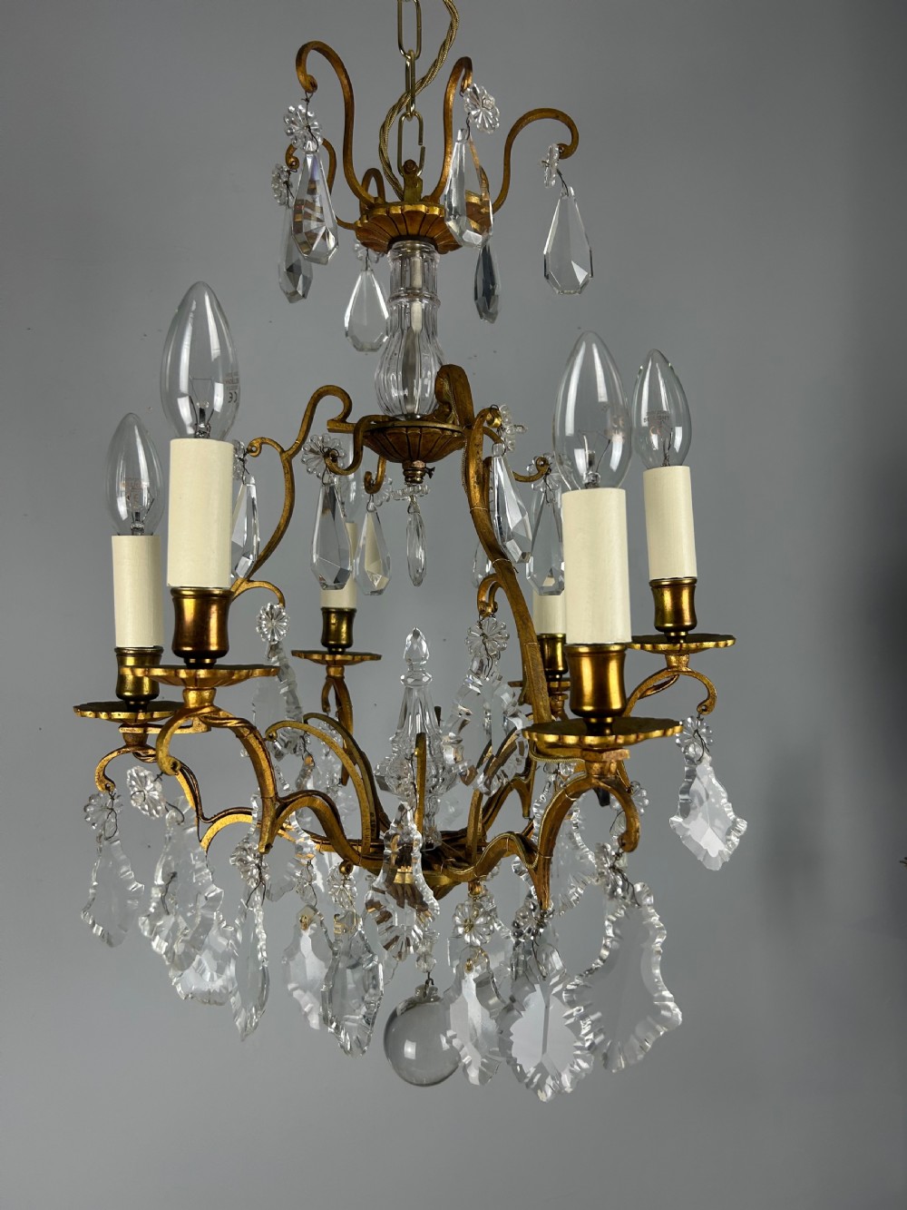 A French brass six-light chandelier featuring curved scroll arms, candle sleeves, and suspended faceted crystal glass droplets. View X