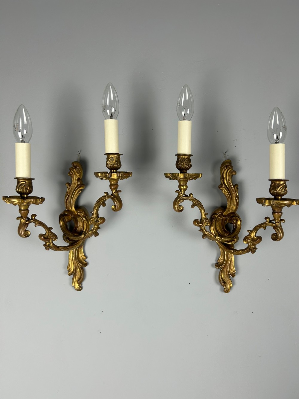 A pair of ornate French gilt bronze wall sconces with twin scrolling foliage arms and white candle-style fittings.