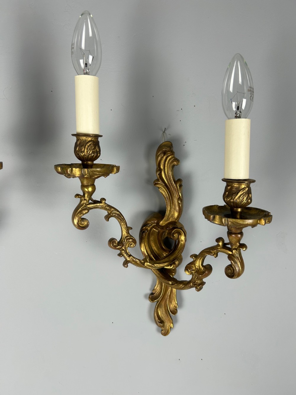 A pair of ornate French gilt bronze wall sconces with twin scrolling foliage arms and white candle-style fittings. View IV
