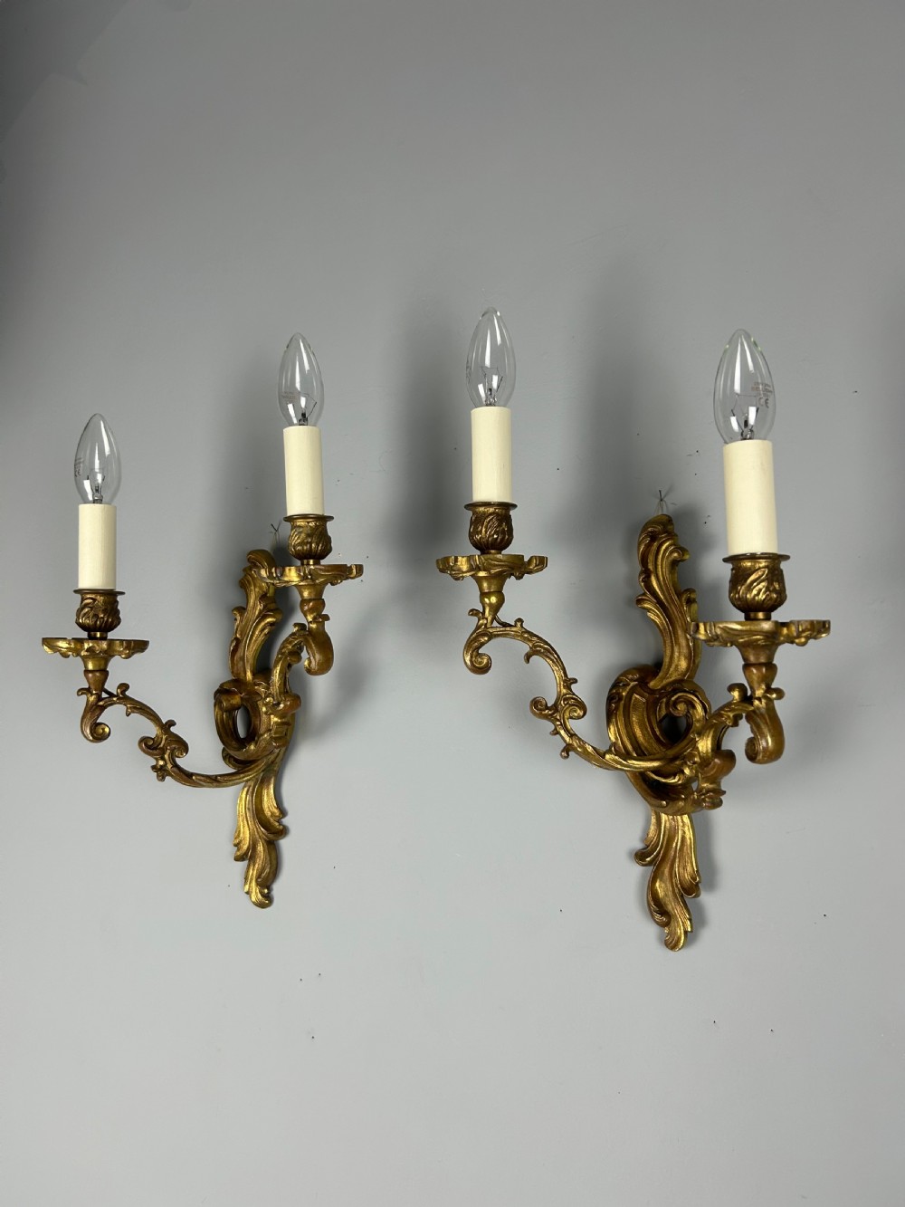 A pair of ornate French gilt bronze wall sconces with twin scrolling foliage arms and white candle-style fittings. View VI