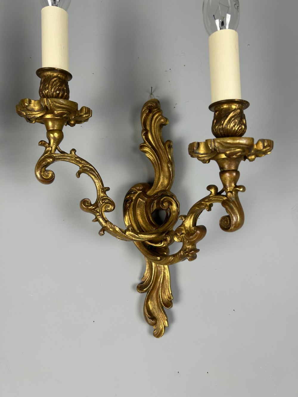 A pair of ornate French gilt bronze wall sconces with twin scrolling foliage arms and white candle-style fittings. View VII