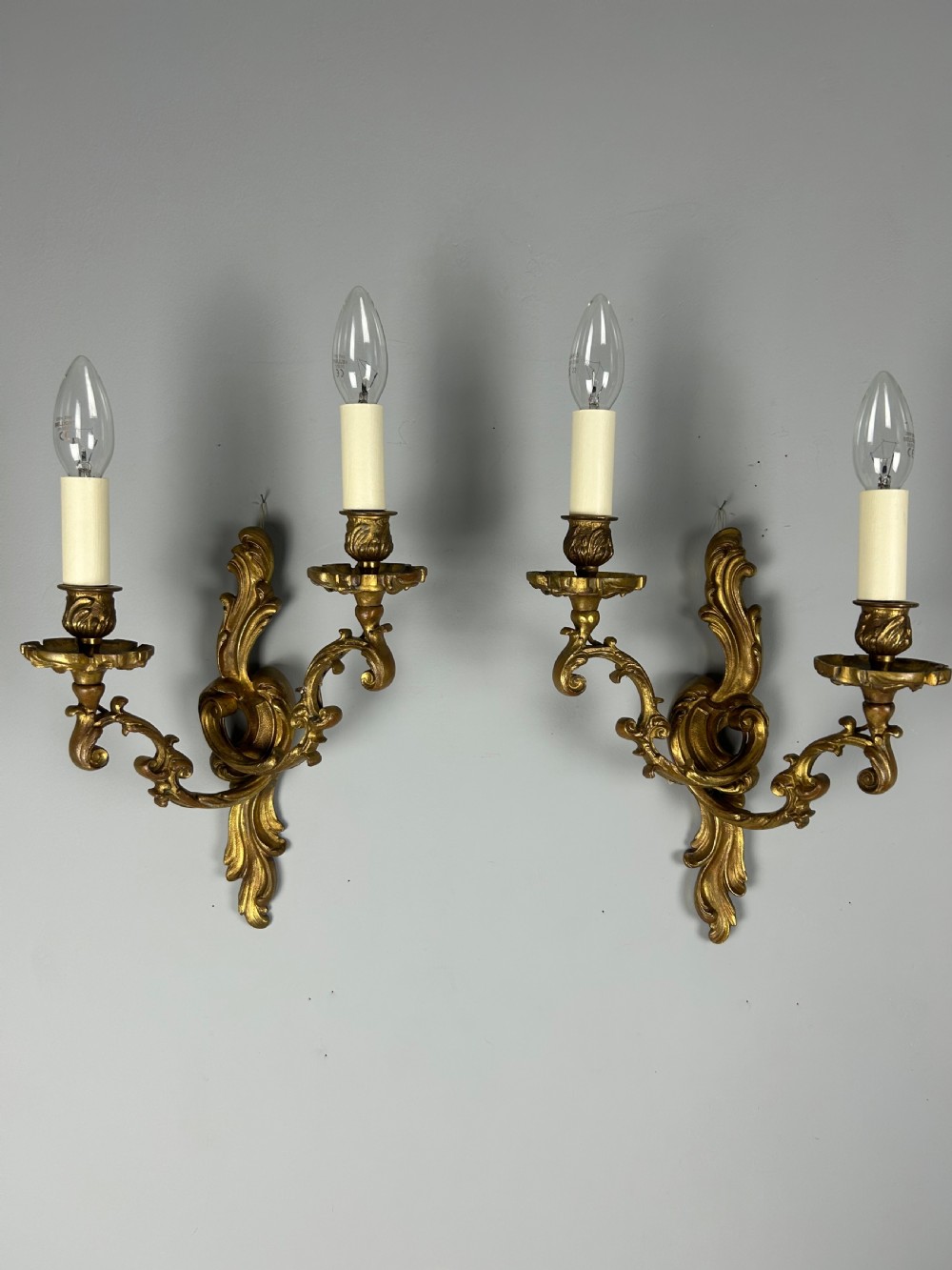 A pair of ornate French gilt bronze wall sconces with twin scrolling foliage arms and white candle-style fittings. View VIII