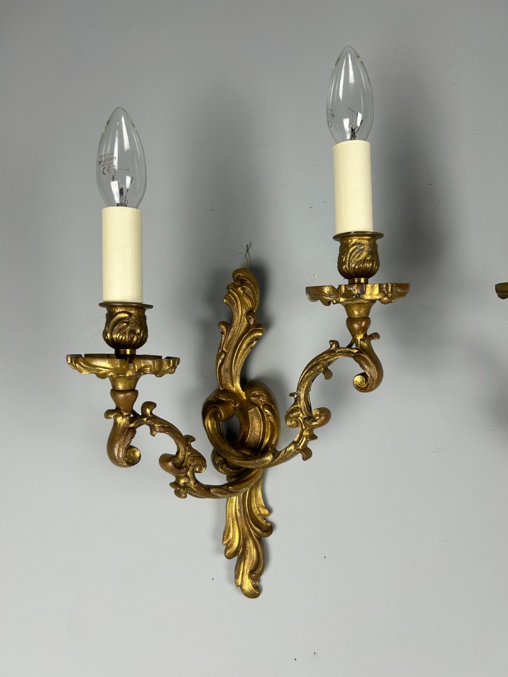 A pair of ornate French gilt bronze wall sconces with twin scrolling foliage arms and white candle-style fittings. View IX