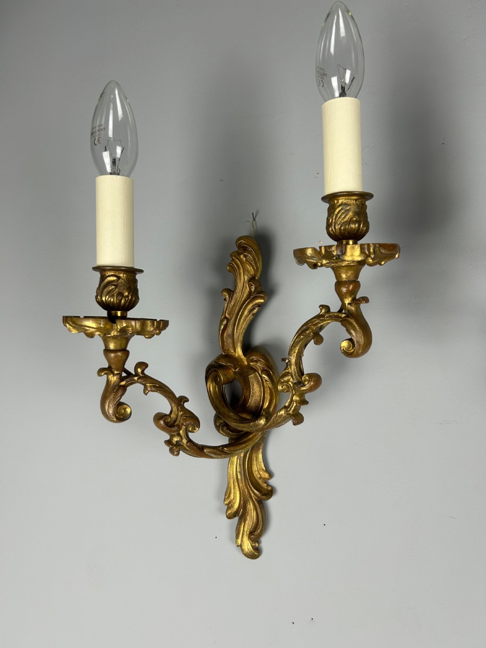 A pair of ornate French gilt bronze wall sconces with twin scrolling foliage arms and white candle-style fittings. View X