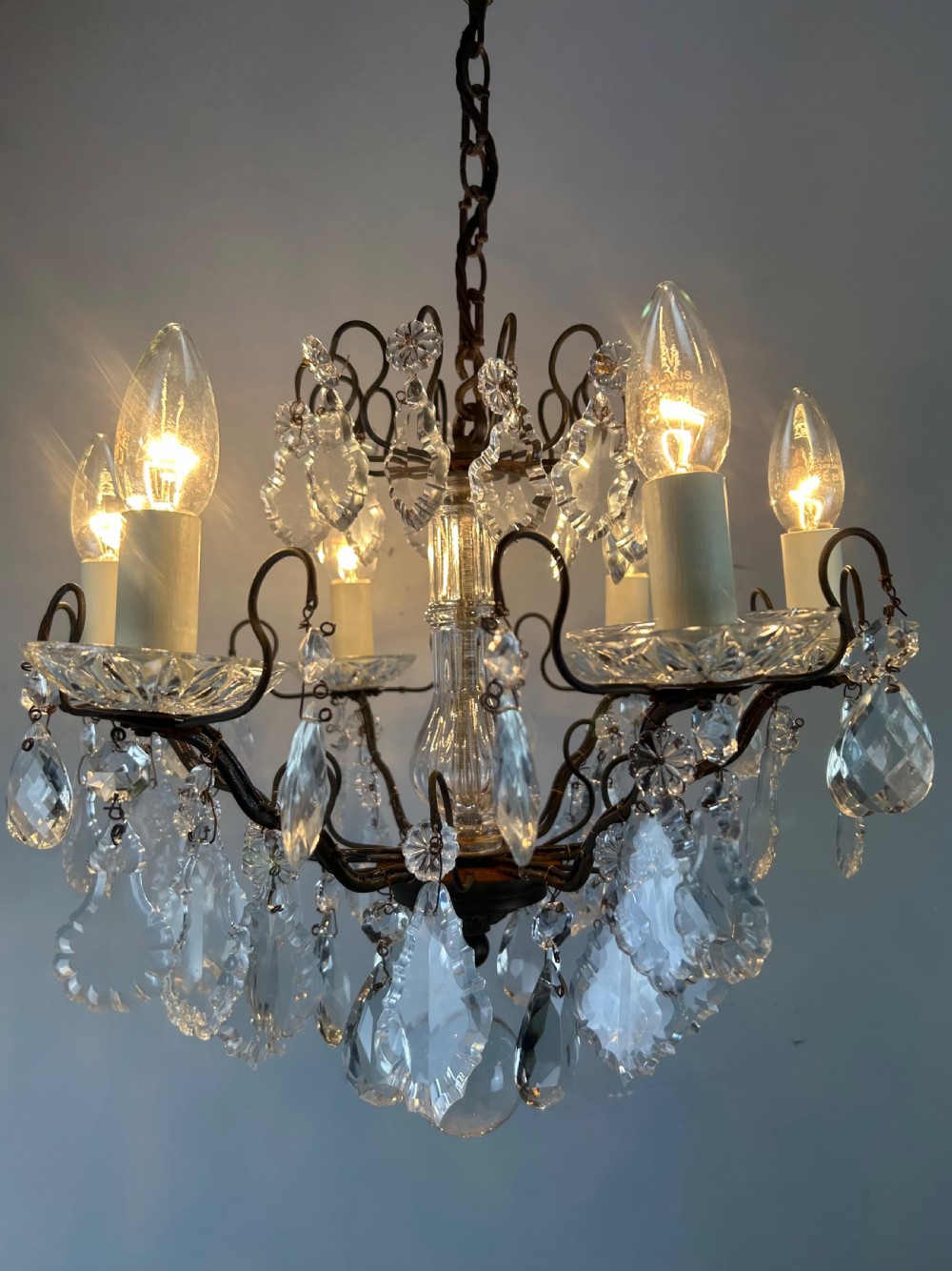An antique French six-light chandelier crafted from dark patinated brass scrollwork and adorned with faceted glass crystal drops and candle-effect fittings. View V